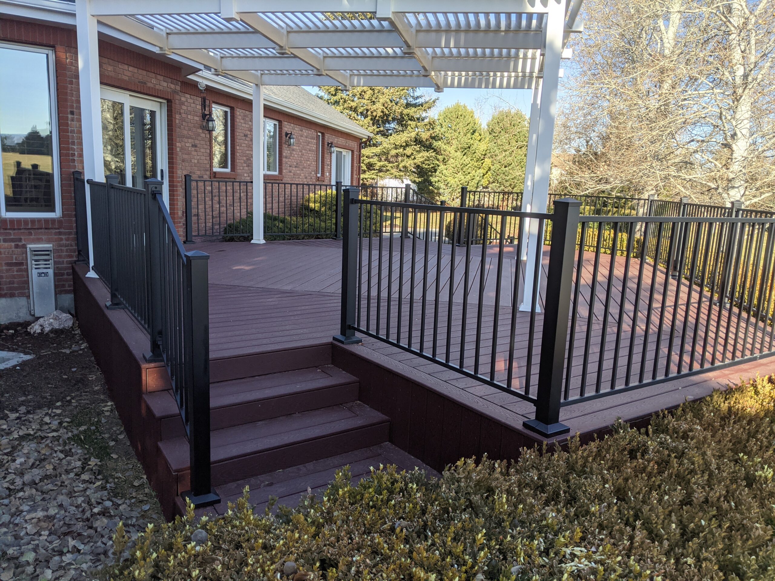 This elegant project by DeckPro LLC features a rich red composite deck that perfectly complements the home's brickwork. The white vinyl pergola provides beautiful filtered shade, while the black metal railing adds a modern touch to this backyard retreat.