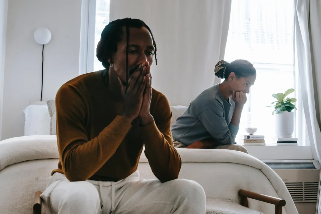 A man and a woman are sitting on a bed, with both appearing to be in distress. The man is in the foreground, wearing a rust-colored long-sleeved shirt and white sweatpants, with his hands clasped in front of his face. The woman is in the background, to the right of the man, wearing a blue sweatshirt and resting her chin on her hand as she looks away. The room is sparsely decorated with a white wall, a white lamp on the wall, and a window with white curtains. A potted plant sits on the windowsill, next to a stack of books.
