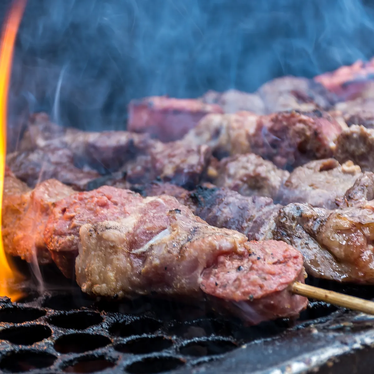A close-up shot of raw meat skewers grilling over an open flame, with smoke rising from the meat and a fire visible on the left side of the image. The meat appears to be beef or lamb, cut into cubes and threaded onto wooden skewers. The grill grate is visible below the skewers, showing a dark, textured surface.