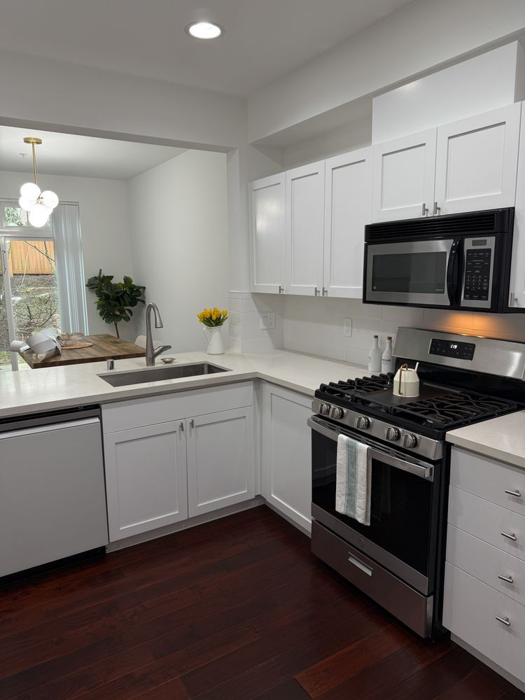 A bright, modern kitchen featuring white cabinets, stainless steel appliances, clean countertops, and dark wood flooring, showcasing a tidy, well-organized space with natural light and minimalist interior design elements.