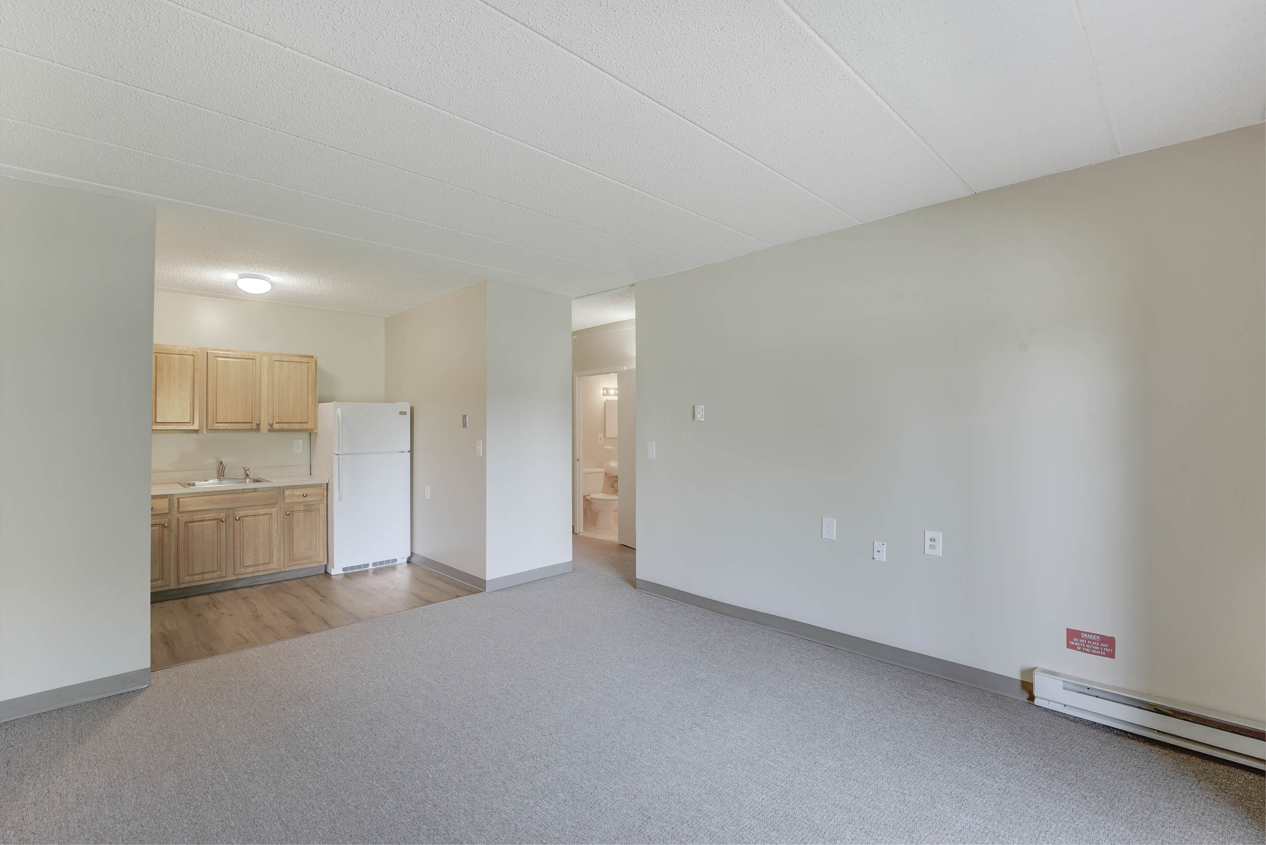 A large empty room with a kitchen area visible through the doorway.