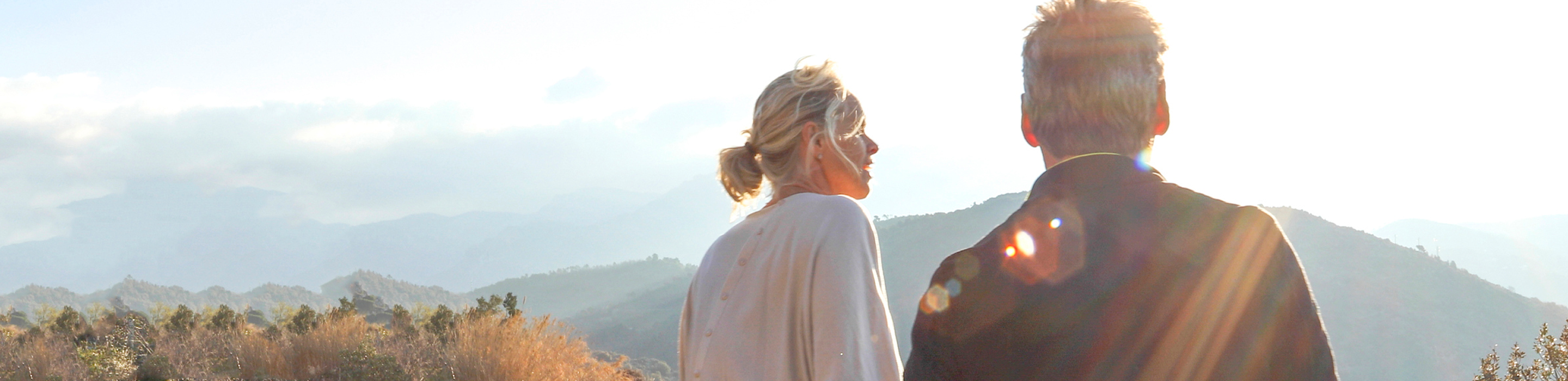 A man and woman sitting on a hill taking in the view of the mountains on a sunny day.