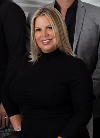 A woman with blonde hair and a black turtleneck smiles in a professional portrait. She is seated and appears to be part of a group photo with other individuals, one of whom is wearing a grey blazer. The background is neutral and the lighting is soft.