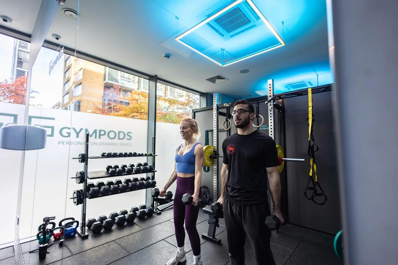 Two people lifting dumbbells in a private Strength POD at GYMPODS Putney with squat rack and dumbbell rack behind them.
