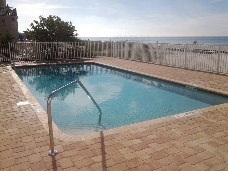 Rectangular beachfront swimming pool with light blue water, stone pavers, metal handrails, and ocean views beyond the fence.