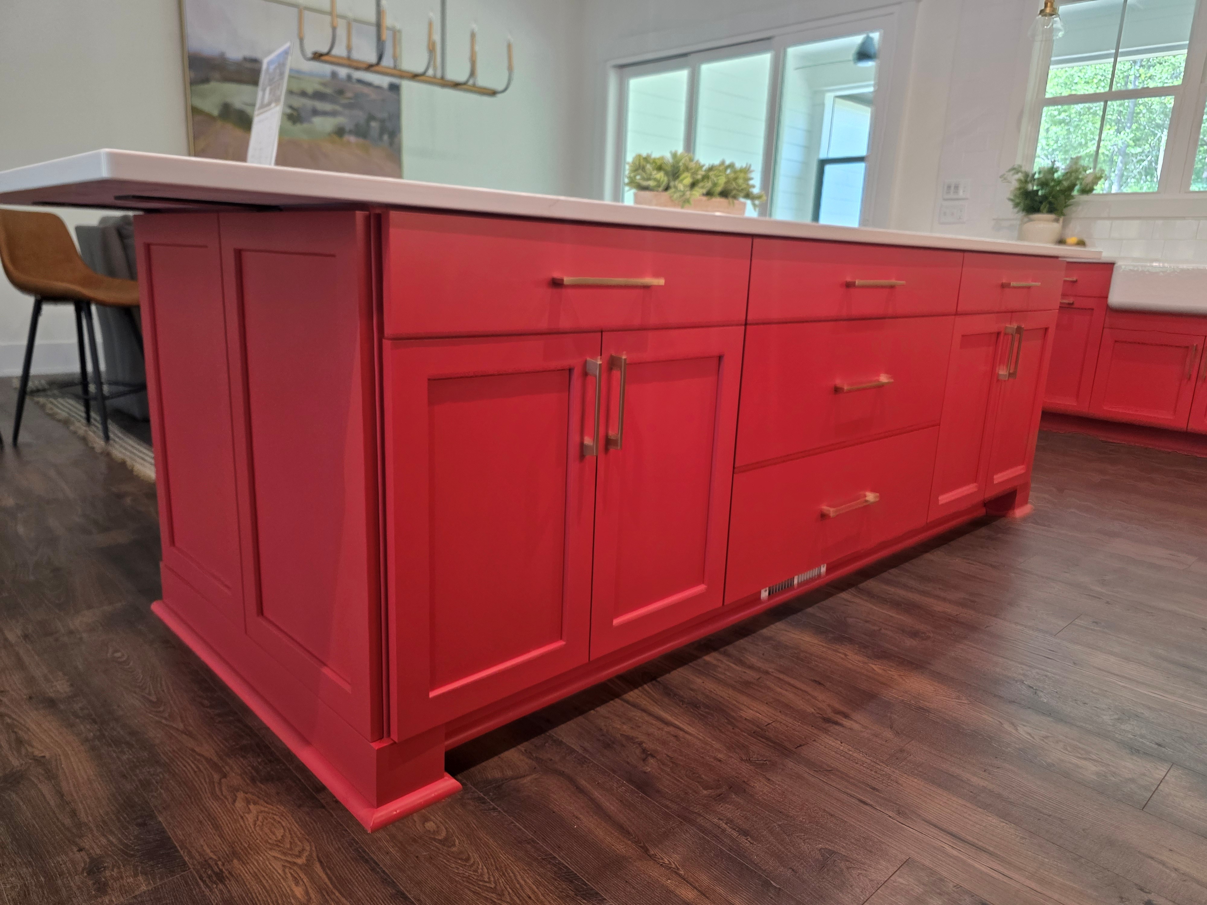 Side view of a red kitchen island featuring gold cabinet hardware, shaker-style doors, and a white quartz countertop, with seating and pendant lighting in the background.