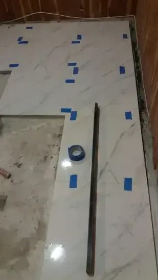 A bathroom floor under construction shows large, white marble-look tiles being laid. Blue painter's tape marks tile placement and spacing. A metal edge and a roll of tape are on the partially tiled surface.