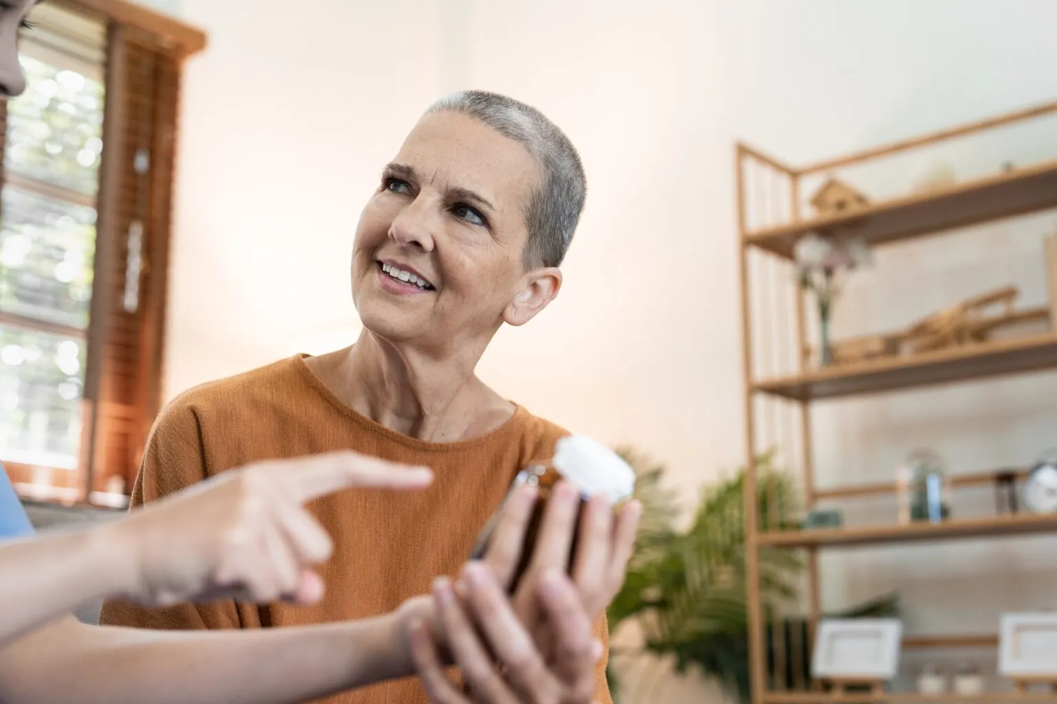 A close-up shot of an older woman with short gray hair, wearing an orange shirt, and a younger person's hand pointing to a medicine bottle. The woman is looking away from the camera and smiling, suggesting a conversation or explanation.