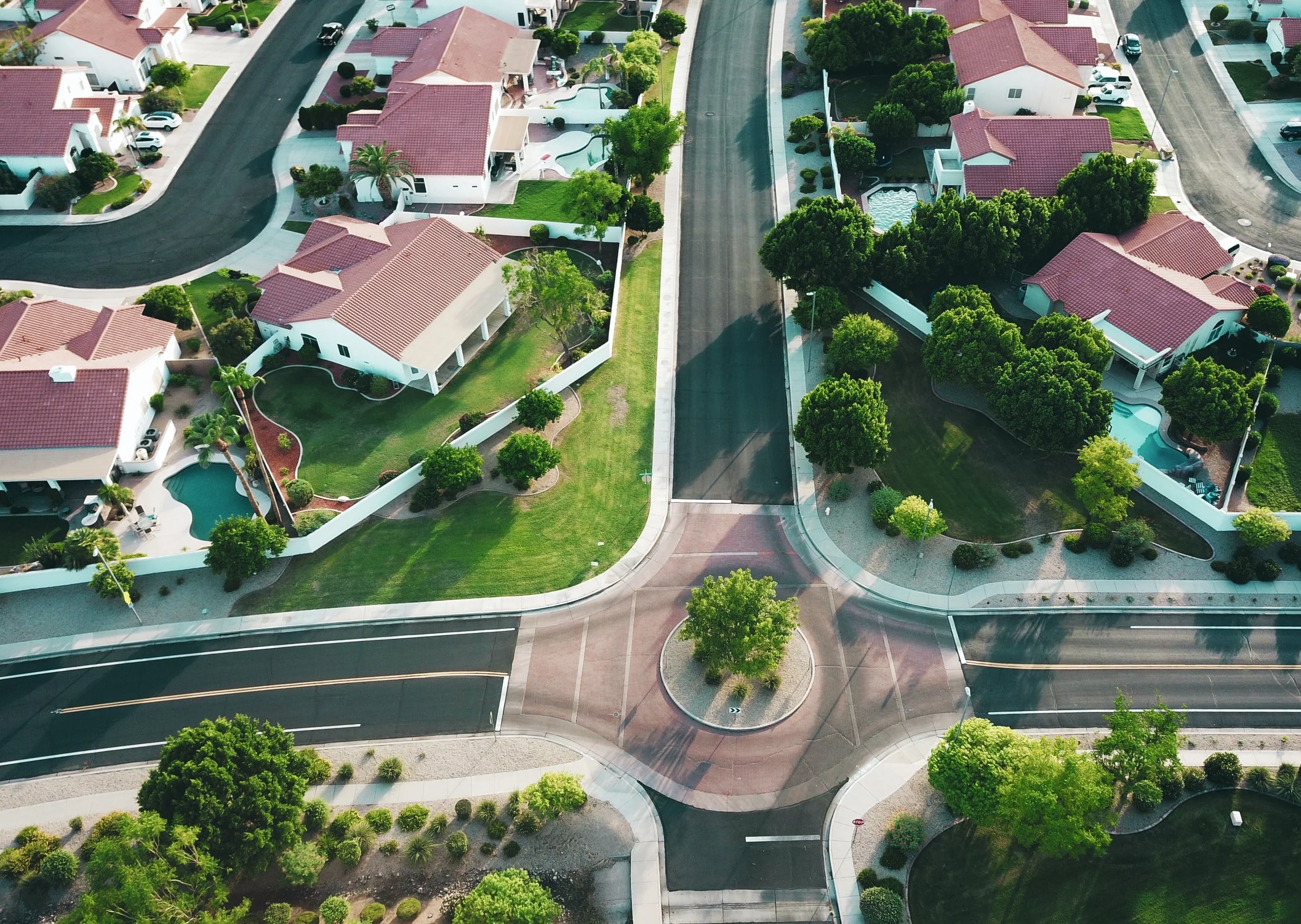 An aerial drone shot captures a suburban neighborhood with well-maintained houses, green lawns, and palm trees. The houses feature red-tiled roofs and white walls. The shot also includes roads with traffic markings, a roundabout with a tree in the center, and several parked cars. Some houses have swimming pools in their backyards.
