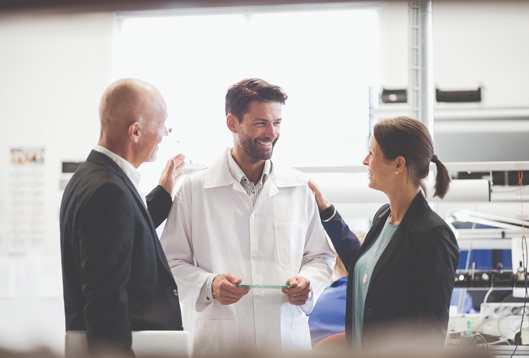 Three people in a lab setting, two in business attire congratulating a smiling man in a lab coat, suggesting teamwork and success.