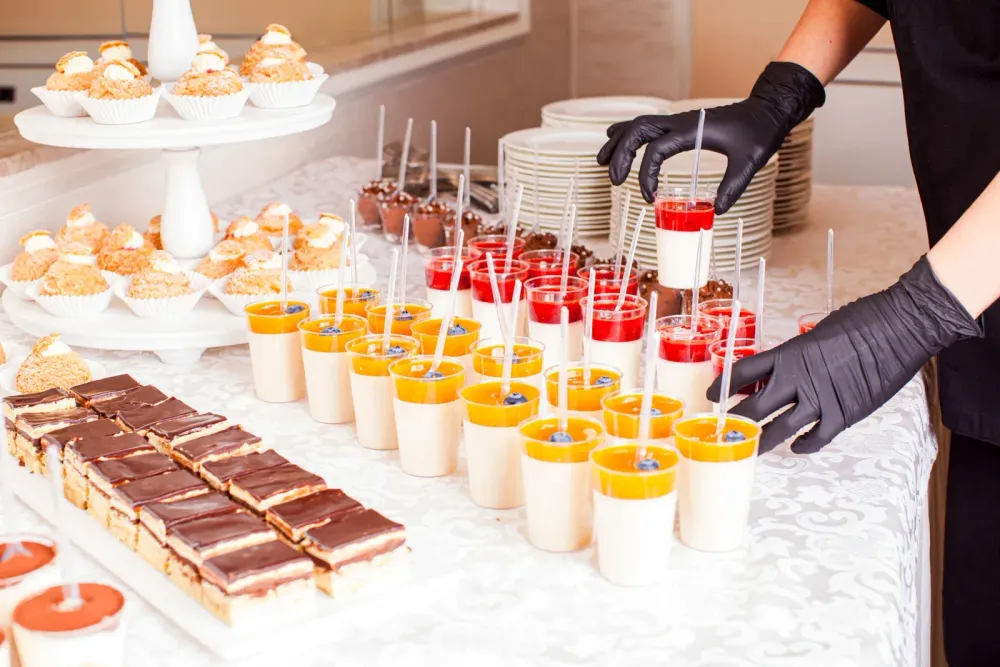 Catering setup featuring assorted desserts arranged neatly on a table, including layered pudding cups, chocolate pastries, and cupcakes. A person wearing gloves organizes the treats, highlighting professional food presentation and preparation for events or gatherings.