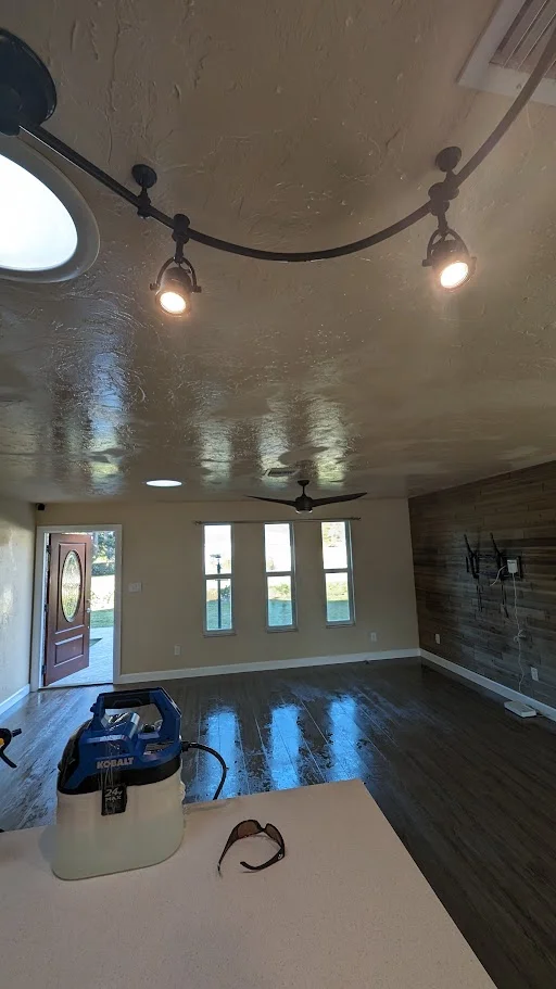 A wide-angle shot of an interior space with a shiny, reflective floor, showcasing a track lighting system on the ceiling, a ceiling fan, a modern wooden accent wall with wall-mounted televisions, and a doorway leading outside. In the foreground, a blue and white portable cleaner and a pair of sunglasses rest on a white surface.