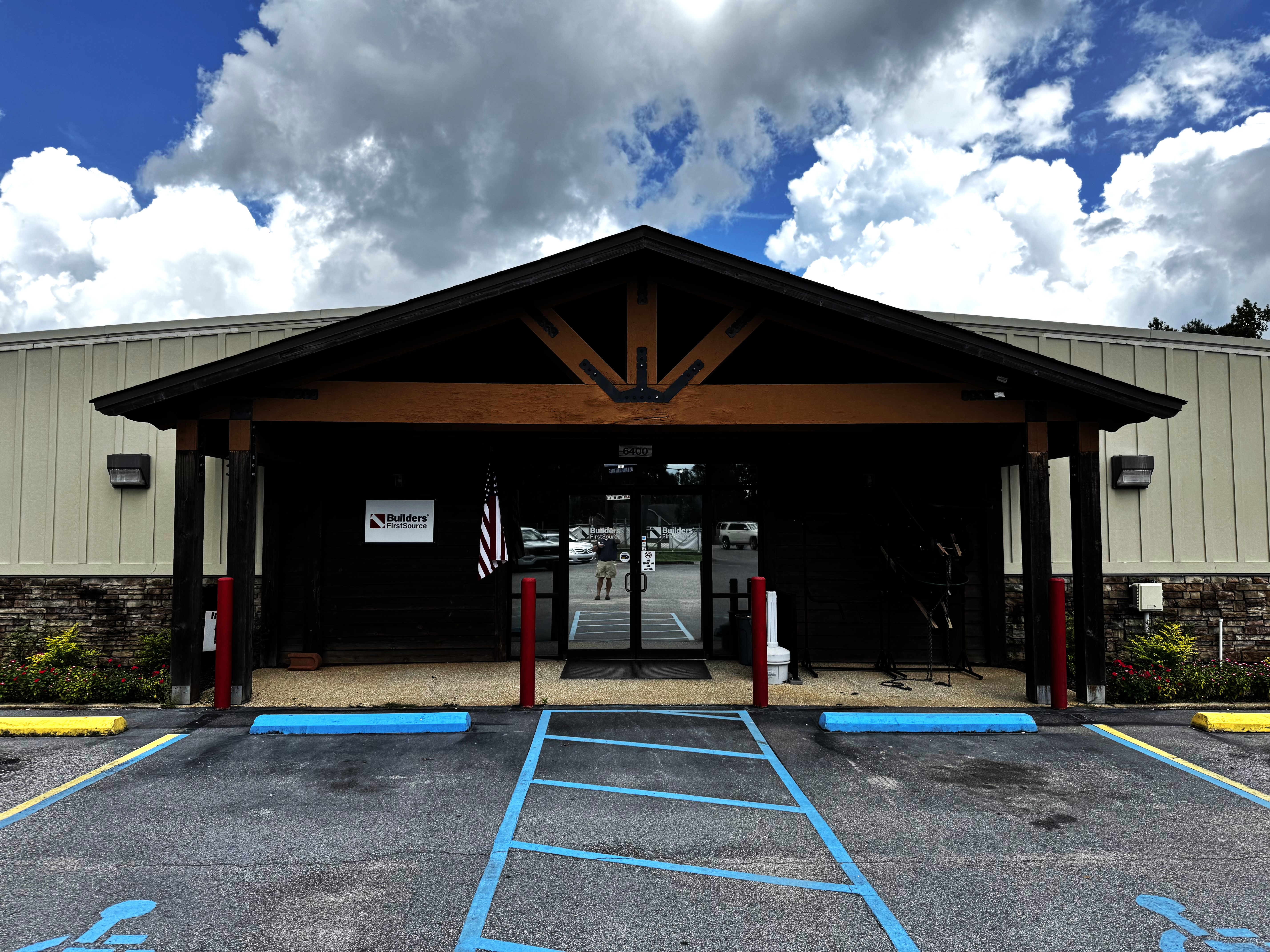 The entrance of a Builders FirstSource facility featuring a rustic wood and metal design with exposed beams. The front includes accessible parking spaces, a stone facade, and an American flag displayed by the glass doors under a partly cloudy sky.