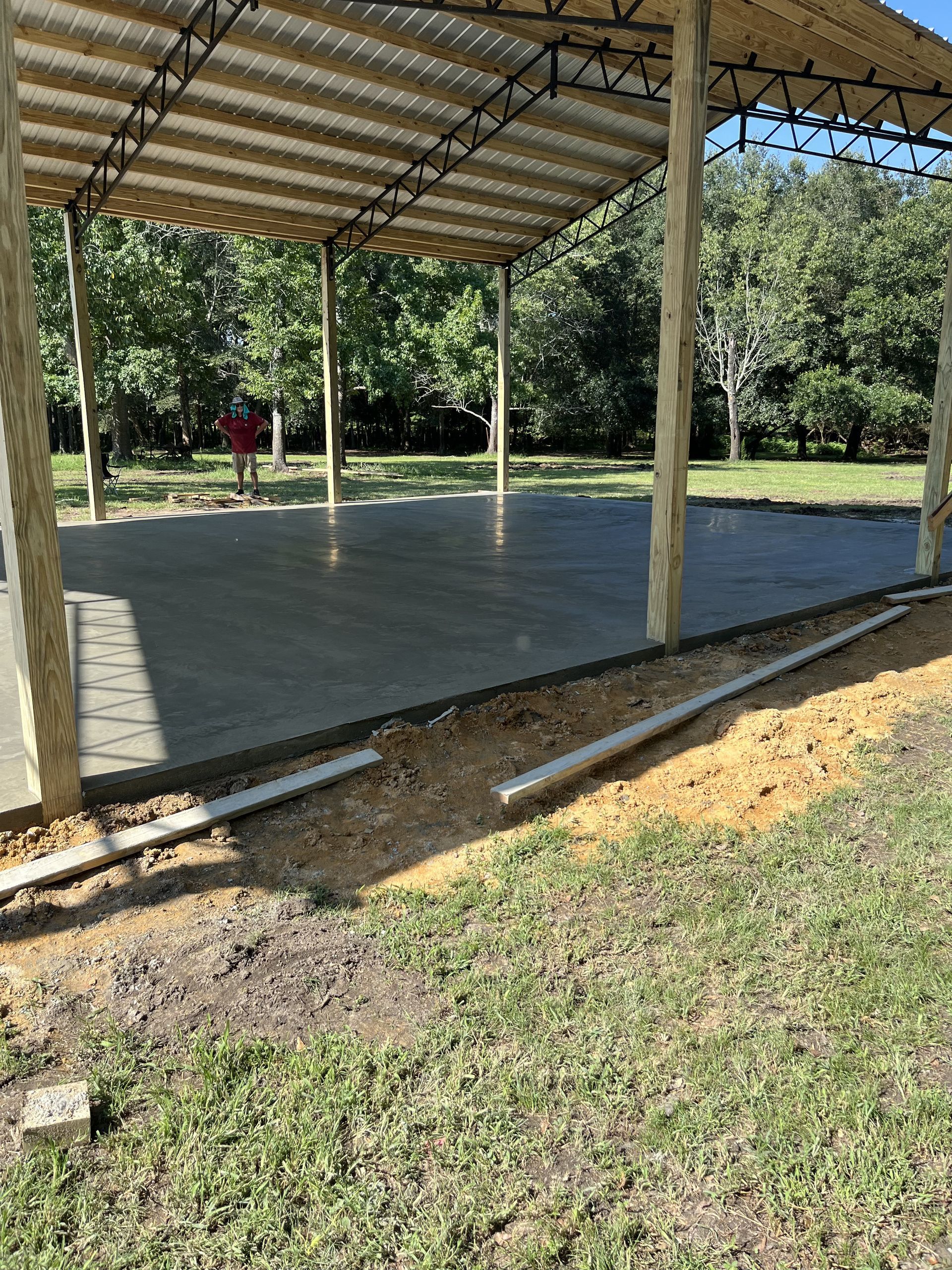 Freshly poured concrete slab beneath open metal roof structure supported by wooden posts and steel trusses, surrounded by grassy yard, with construction boards and worker visible in background.
