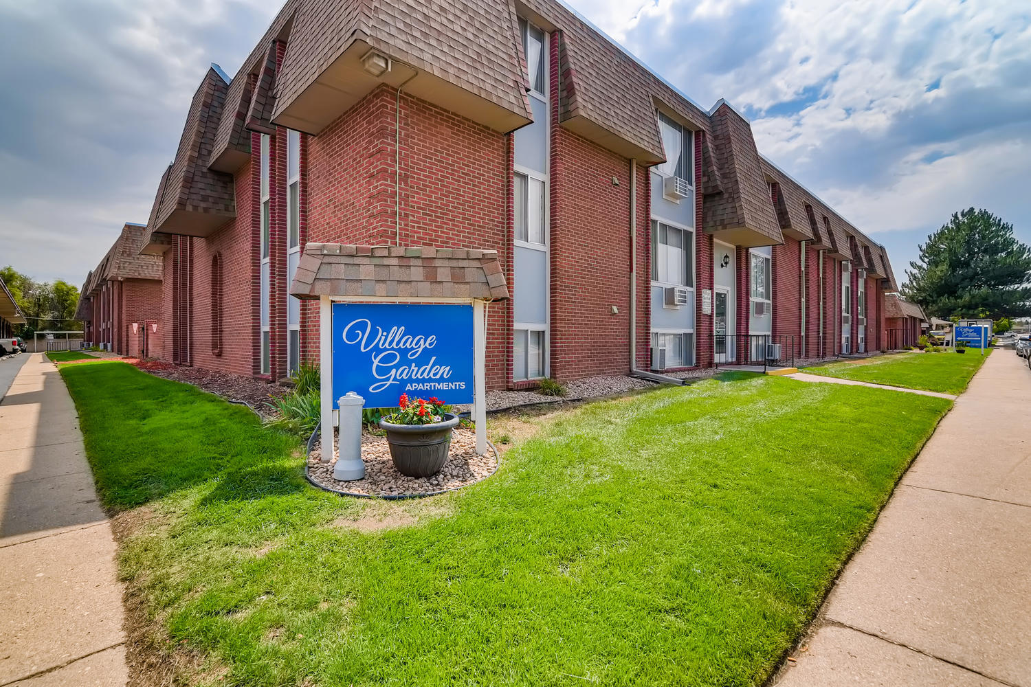 Village Garden blue monument sign with grass, sidewalk, and brick and siding building exterior.