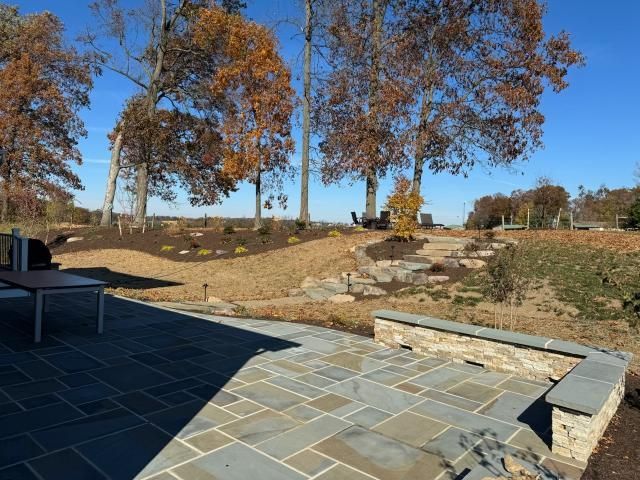 Backyard patio with stone seating wall and surrounding landscape including trees and sloped terrain. The design showcases integrated hardscaping and landscaping, creating a natural yet structured outdoor environment for relaxation and outdoor use.