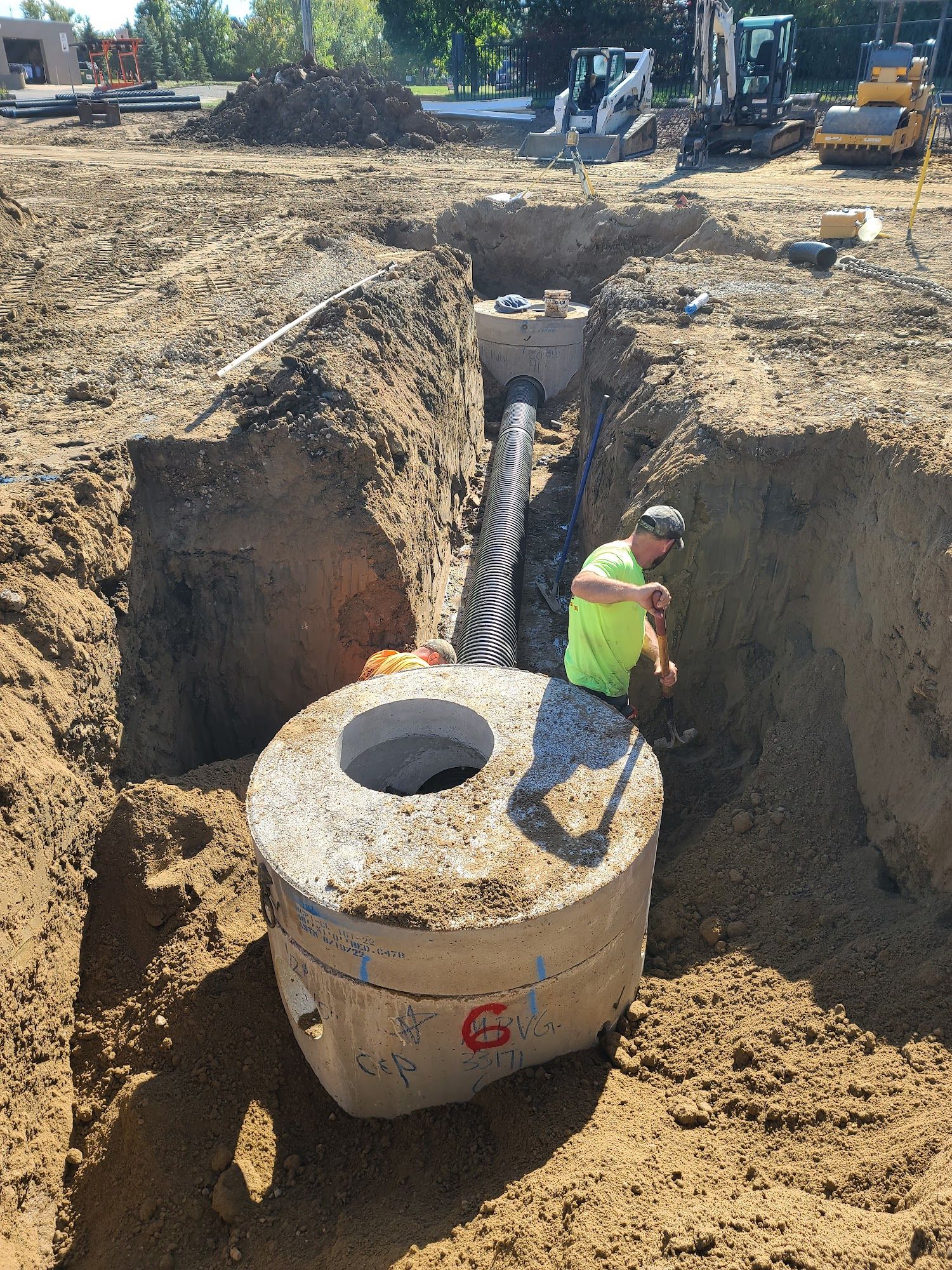 Workers install underground drainage piping between precast structures within a trench. The crew adjusts pipe elevations and connections while preparing the system for stormwater management.