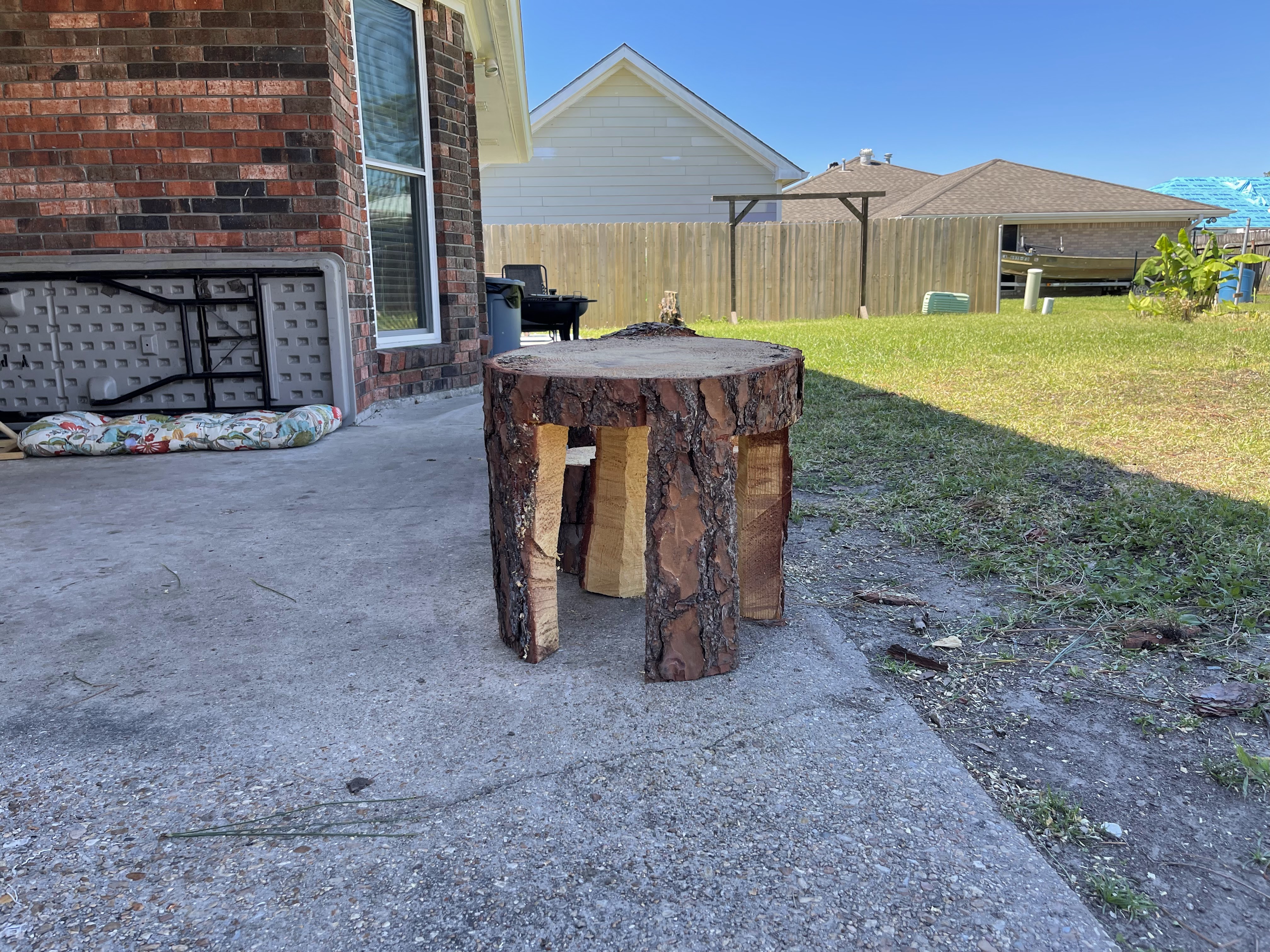 A rustic, log-style stool or small table sits on a concrete patio next to a brick house. The stool is made from a thick tree trunk with bark still attached, and has three wooden supports. In the background, a grassy yard is visible with a wooden fence, a barbecue grill, and houses under a clear blue sky. A colorful cushion is lying on the patio to the left of the stool.