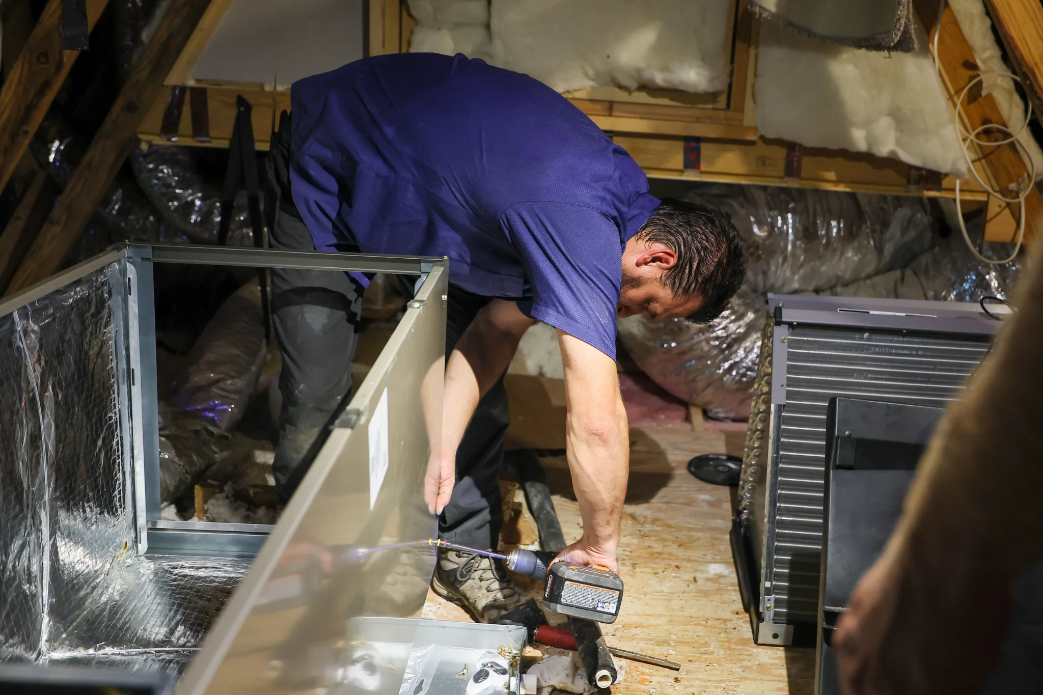 A person in a blue shirt is working in an attic, using a cordless drill to attach a piece of equipme 