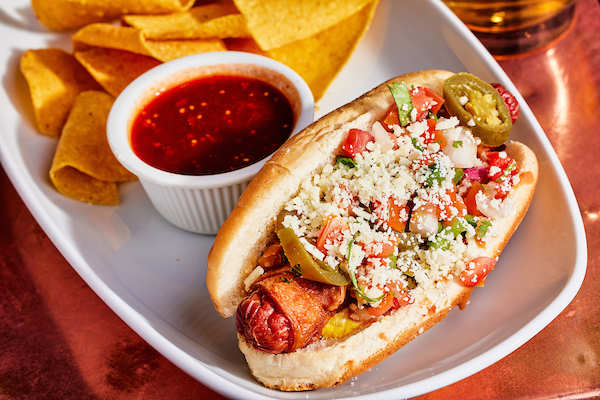 Latin style hot dog topped with jalapenos and cheese with chips and salsa on the side 