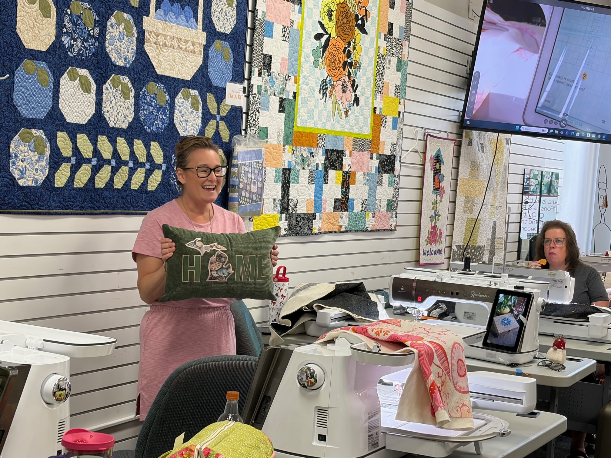 A participant proudly displays her completed HOME pillow during a Decorative Stitch class. This creative project showcases decorative stitching and machine embroidery techniques learned in a fun, hands-on workshop.