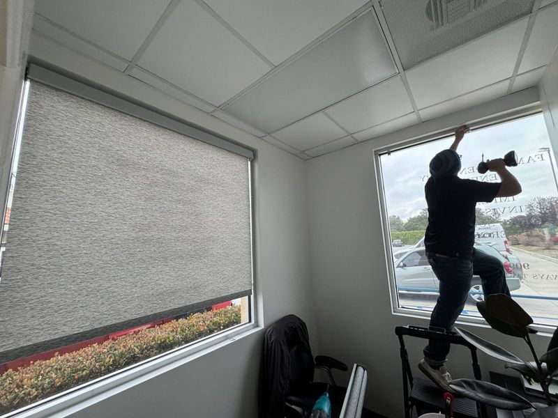 Professional installation of custom roller shades in a commercial office. One window features a sleek, textured gray shade, while a technician carefully mounts the matching hardware for the second pane.