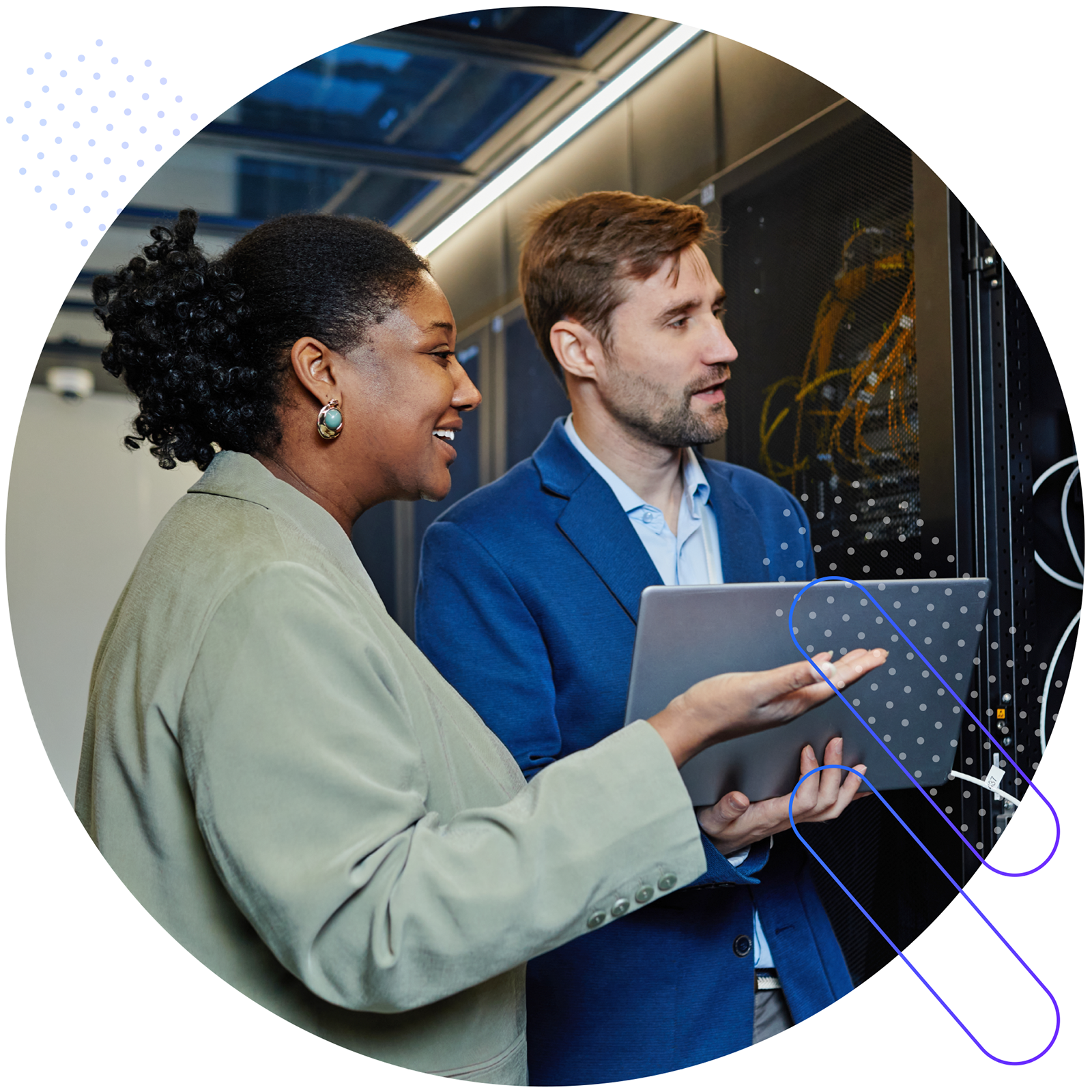 Two professionals, a Black woman and a White man, stand in a server room, reviewing data on a laptop. The woman is wearing a light green blazer and large, turquoise earrings. The man is wearing a blue suit jacket over a light blue dress shirt. They are both looking at the laptop screen, with the woman pointing at it. The server racks and cables are visible in the background.