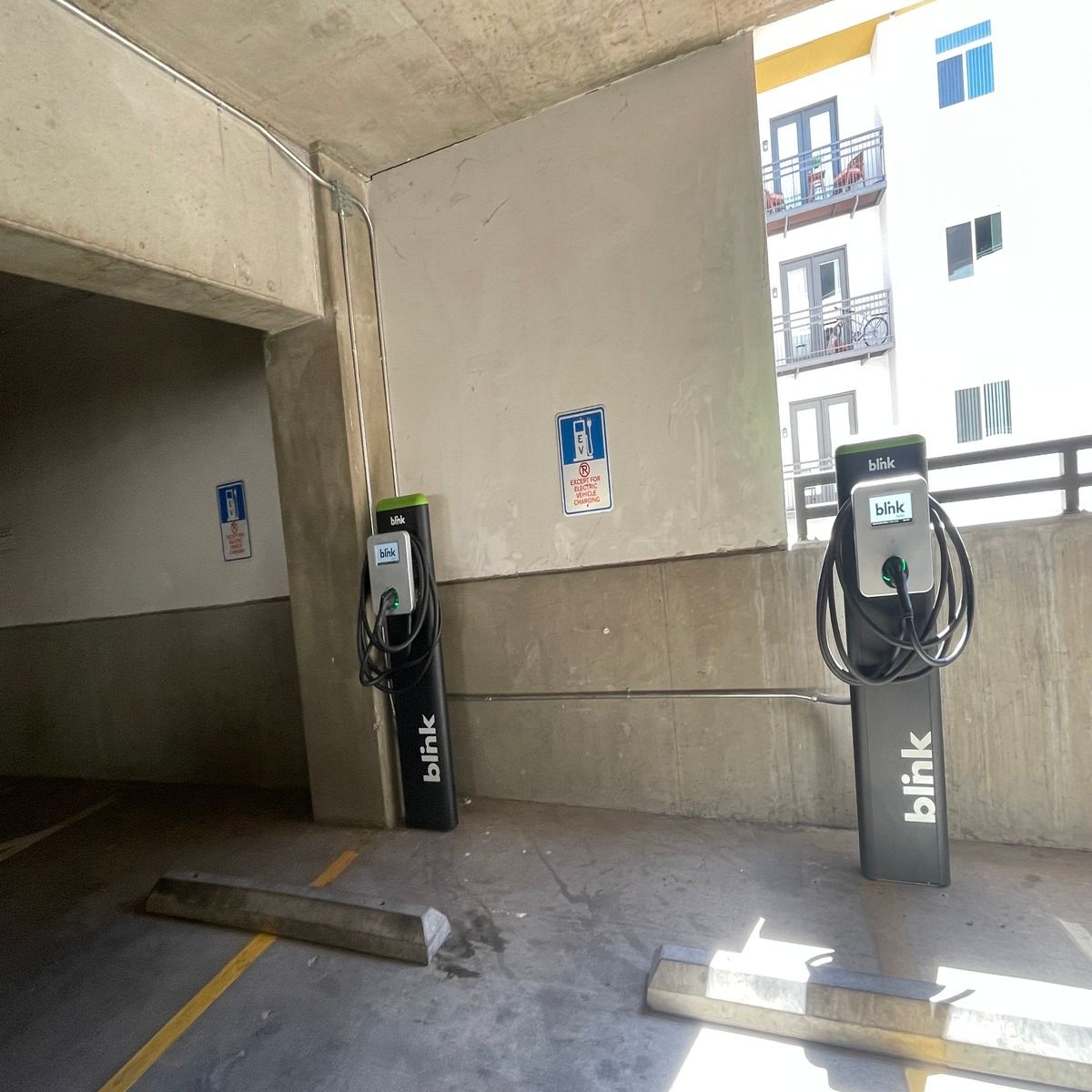 Two Blink electric vehicle charging stations stand in designated parking spaces inside a concrete parking garage, each mounted beneath EV charging signs with coiled cables ready for drivers to use.