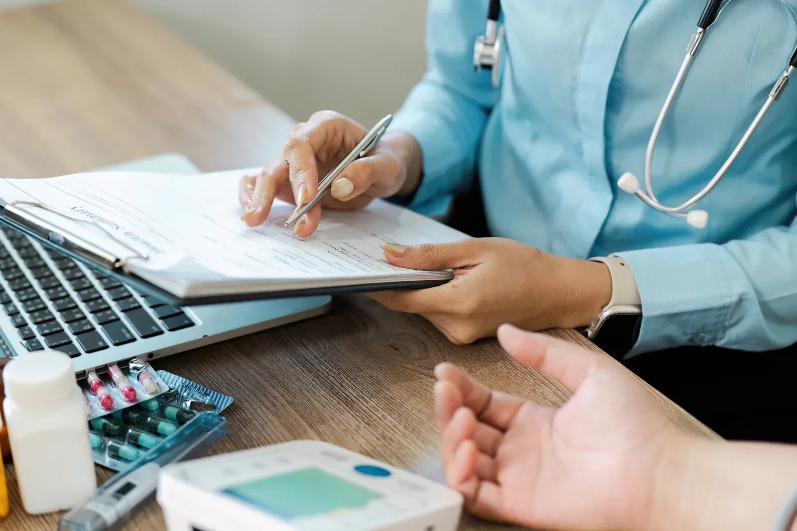 A healthcare professional with a stethoscope reviews and fills out paperwork on a clipboard while consulting a patient, surrounded by medical supplies, pills, and a digital device on a wooden desk.