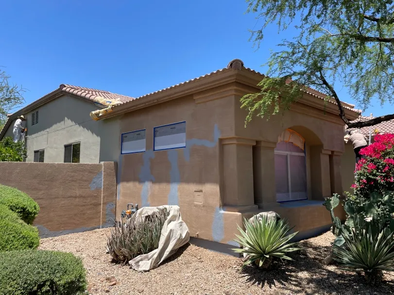 Residential exterior painting project showing stucco home with patched primer areas, masked windows, and workers preparing surfaces, surrounded by desert landscaping, indicating ongoing repainting work under bright, sunny outdoor conditions.
