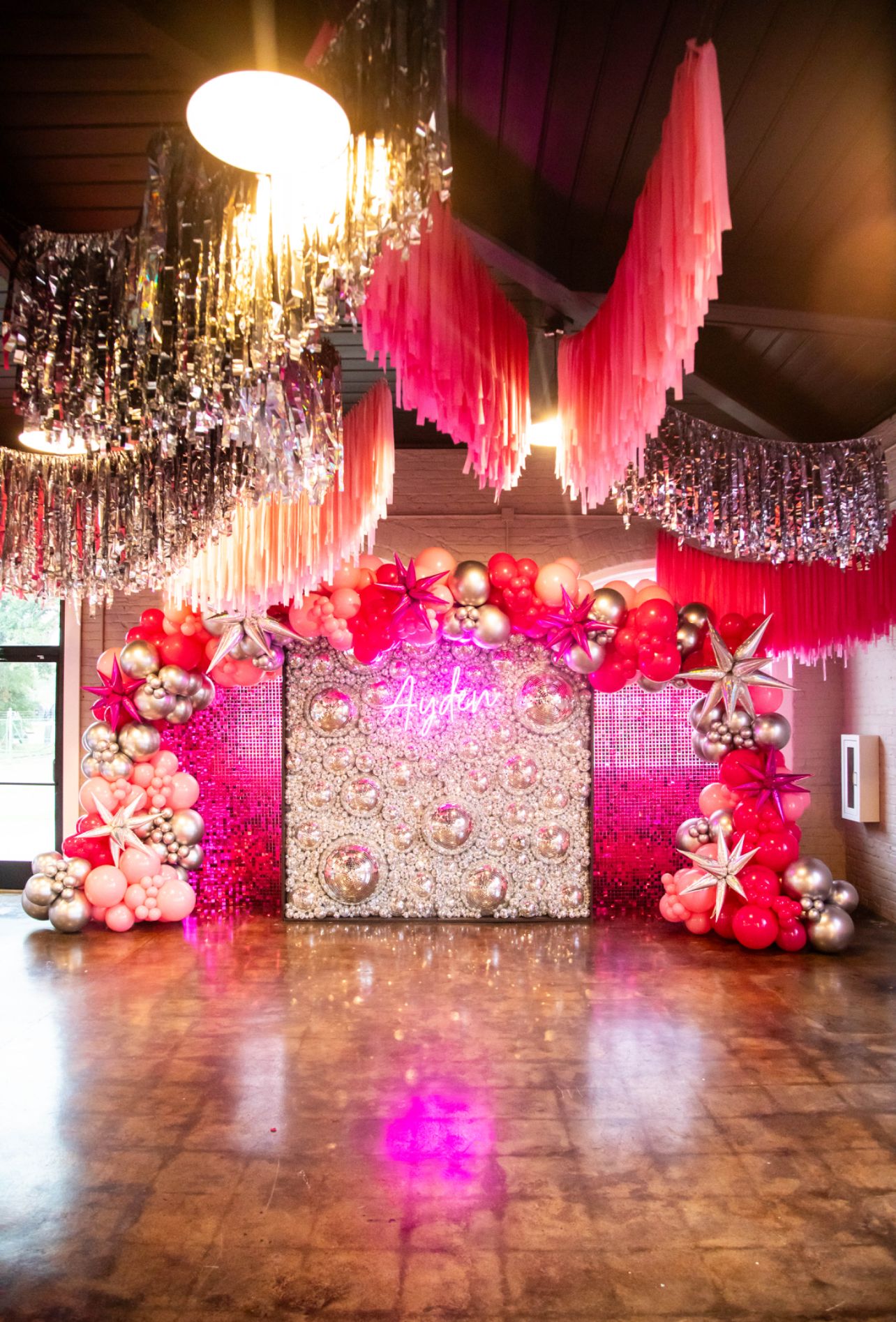 Luxurious party backdrop featuring pink, red, and metallic balloon arch, mirrored floral wall with neon name sign, hanging fringe ceiling d&eacute;cor, and shimmering accents, creating an elegant, glamorous indoor celebration photo setup.