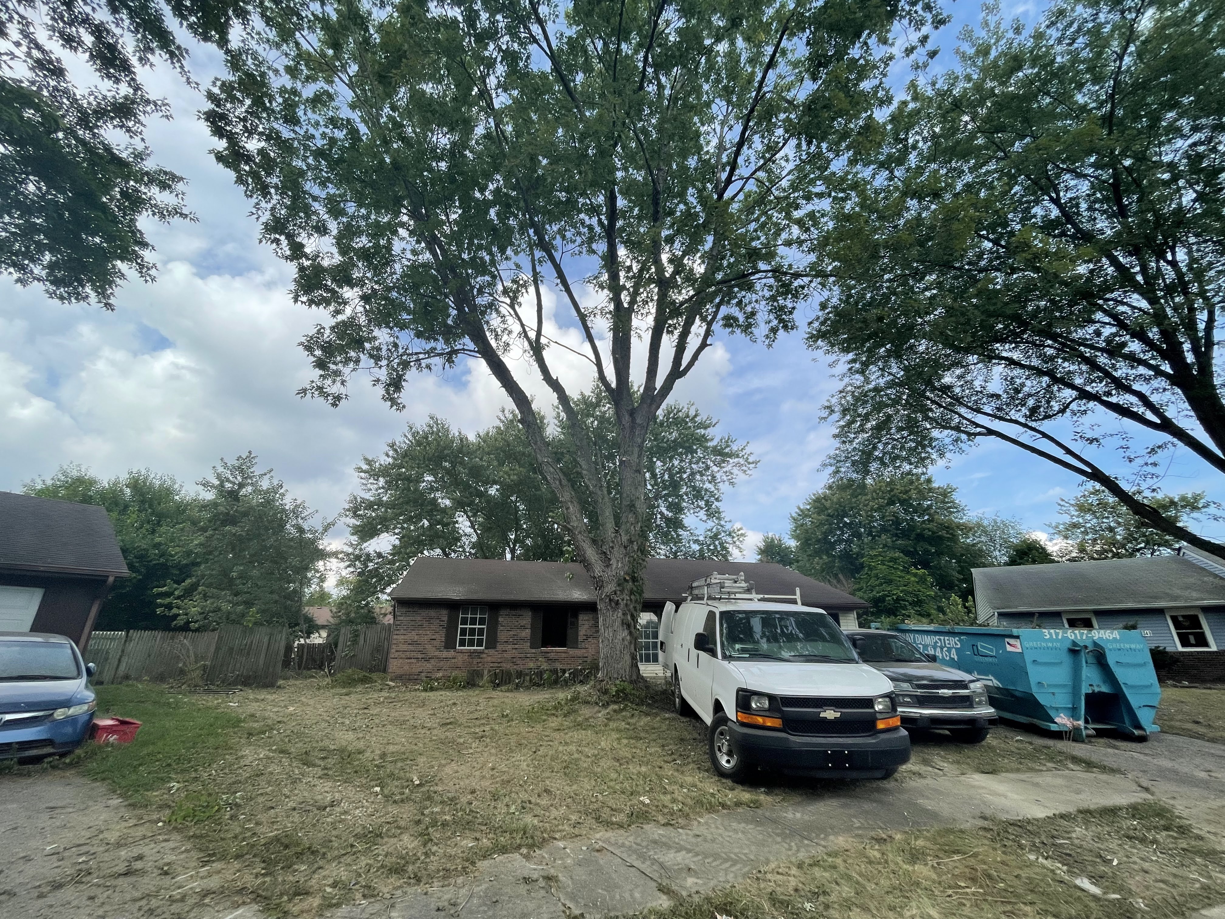 A white van is parked in front of a brick house with a large tree in the yard. Two large blue dumpsters are also visible in the driveway. The sky is partly cloudy.
