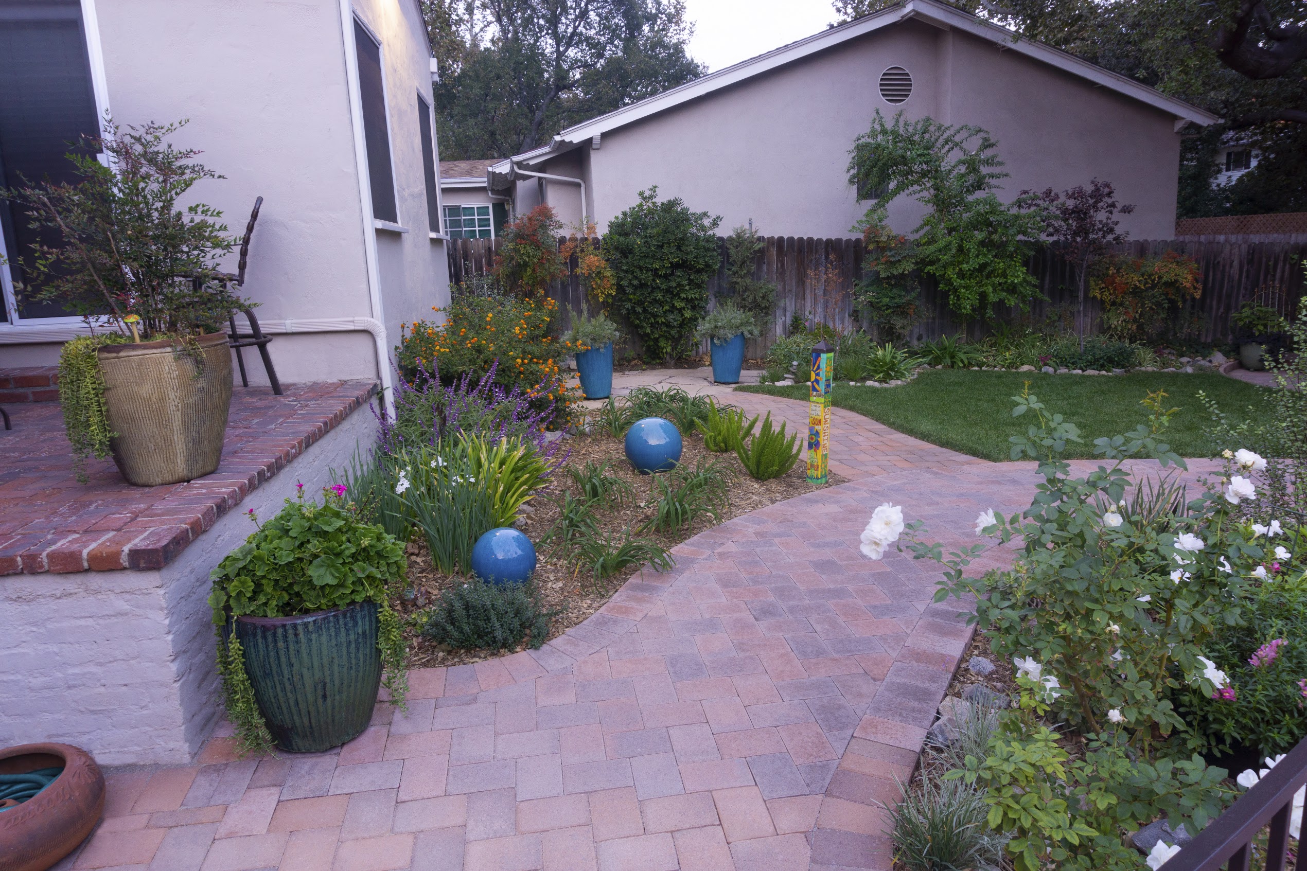 Colorful backyard garden with brick patio, potted plants, flowers, blue gazing balls, and a winding path, creating a peaceful, welcoming mood.