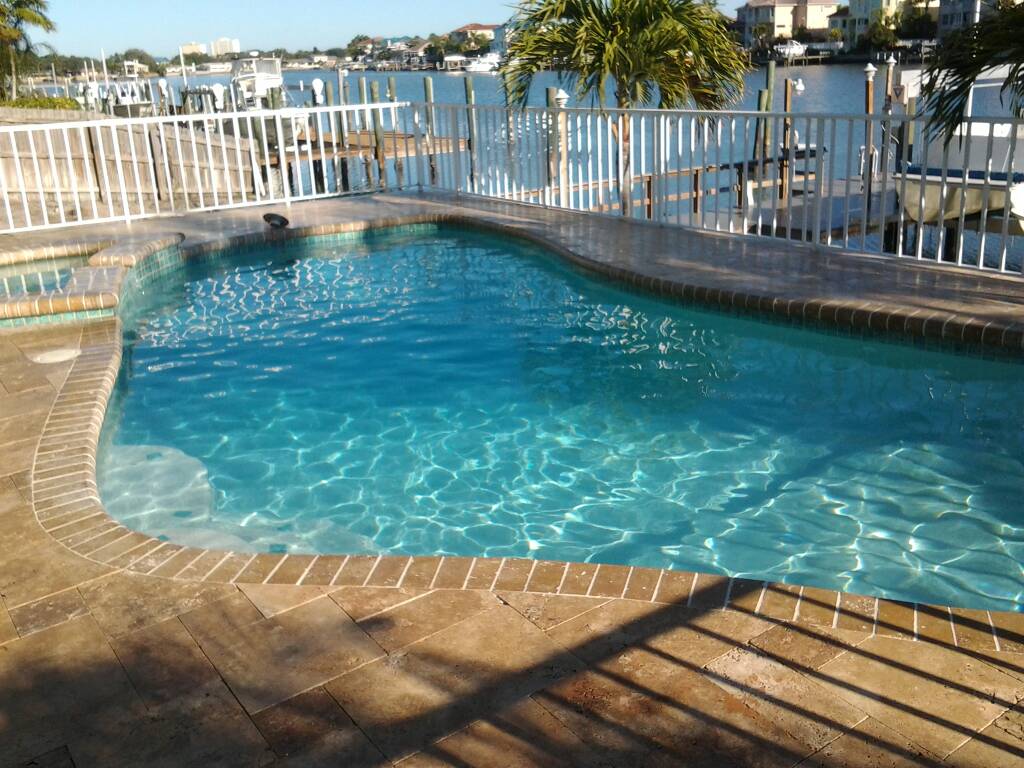 Waterfront backyard swimming pool with bright blue water, brick paver decking, and a white safety fence overlooking the water.