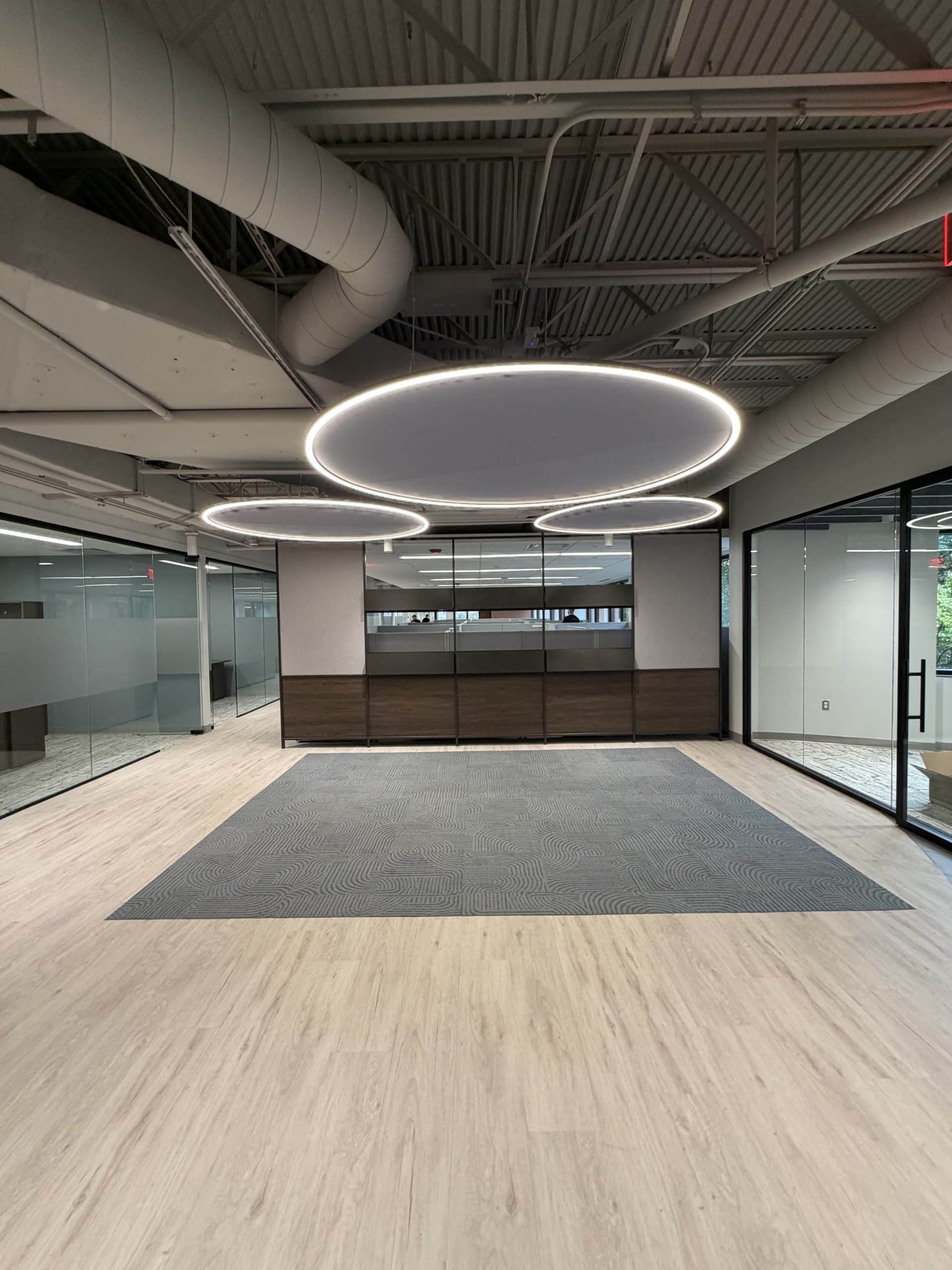 The Toll Brothers Michigan Division Office & Design Studio features a contemporary office interior highlighted by circular suspended lighting, glass partitions, and coordinated flooring. Part of Schonsheck, Inc.’s commercial construction portfolio, this space pairs an exposed ceiling with refined architectural finishes to create an open, professional atmosphere. Clear sightlines and glass-front rooms connect the central area with surrounding workspaces, showcasing the visual appeal and functionality of modern commercial office interiors.
