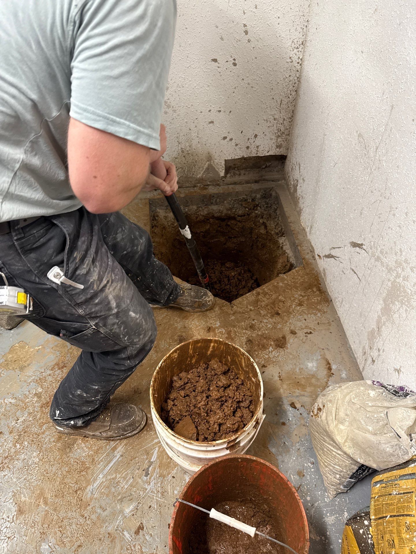 Worker excavates a square opening in a concrete floor, using a long digging tool while wet soil and debris fill buckets nearby, indicating active underground plumbing or drainage access work.