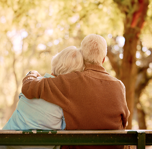 An elderly couple sitting on a bench in a park, as seen from behind.