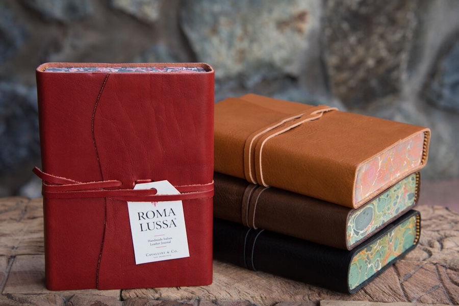 A classic product image by Tabula Rasa Social Stationers, showcasing a collection of Cavallini Roma Lussa Handmade Italian Leather Journals. The foreground features a prominent red journal, standing upright, highlighting its rich leather texture and signature wrap-around tie closure with a product tag. Behind it, a stack of journals in tan, dark brown, and black reveals their beautifully marbled page edges, embodying Cavallini’s timeless charm and vintage-inspired artistry. These high-quality journals offer smooth, durable pages, perfect for sketching, jotting notes, or planning.