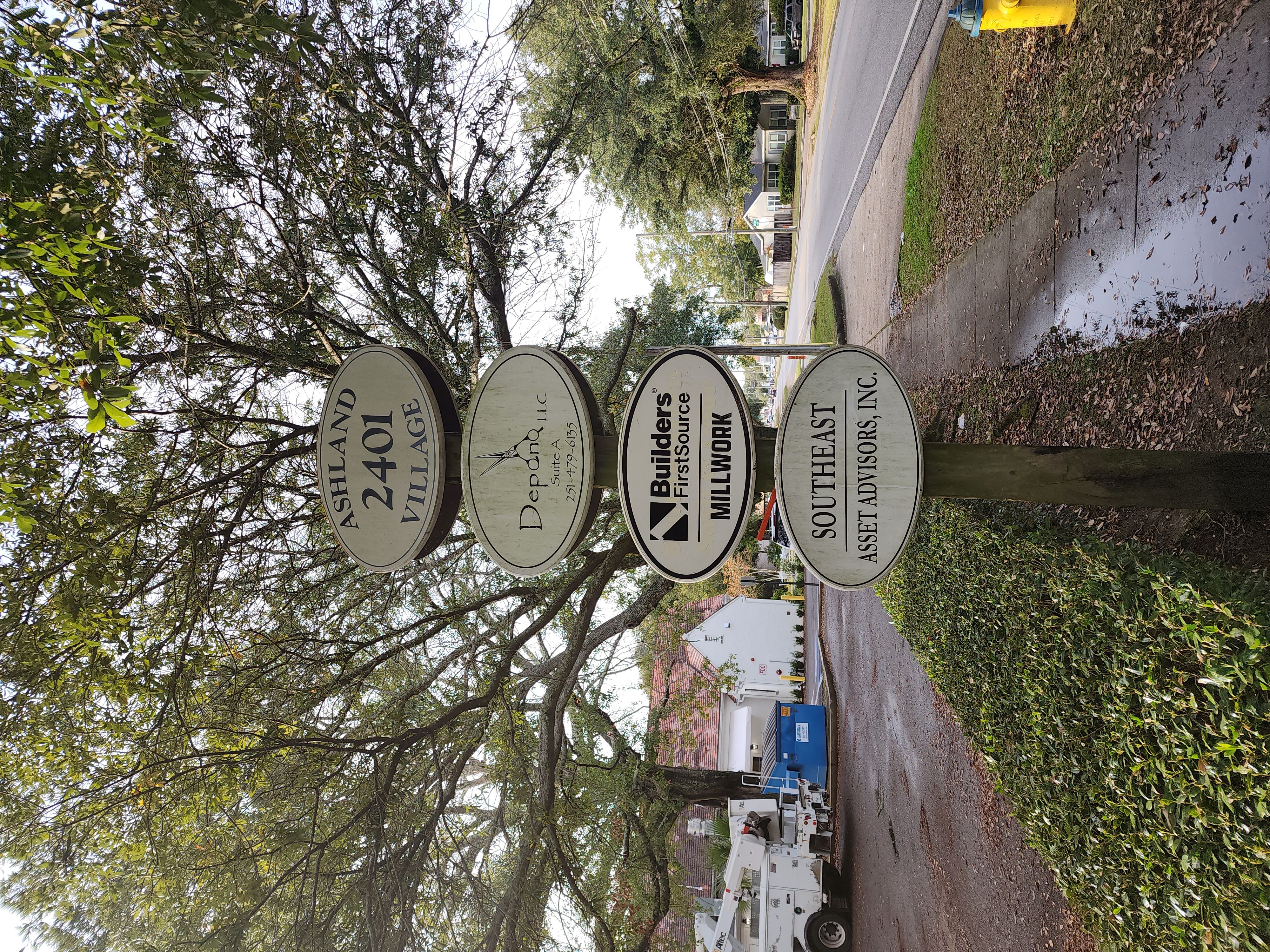 (Directional Signage): A multi-business signpost at 2401 Ashland Village, featuring a sign for Builders FirstSource Millwork. The sign is surrounded by a green hedge, tree canopy, and a neighborhood street in the background.