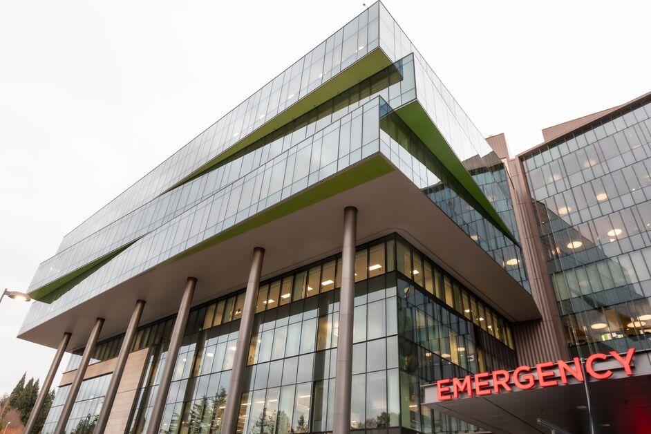 An architectural view of a contemporary glass and steel hospital building featuring cantilevered floors with lime green accents. Tall pillars support the upper levels, and a glowing red "EMERGENCY" sign marks the entrance canopy.