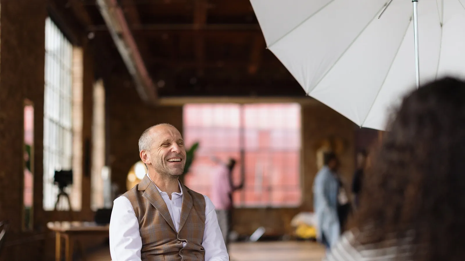 A middle-aged Caucasian man with a warm smile is the focus of this photograph. He is wearing a white dress shirt and a plaid vest, suggesting a semi-formal or business casual attire. The man is positioned in what appears to be a studio or a loft-like space, characterized by exposed brick walls, large windows, and industrial-style lighting. A white umbrella, likely used for photography lighting, is visible in the upper right corner, partially obscuring a person with dark, curly hair in the foreground. In the background, other individuals and studio equipment, including a camera on a tripod, can be seen, indicating a professional photoshoot or filming environment. The overall impression is one of a candid, joyful moment captured during a professional setting.