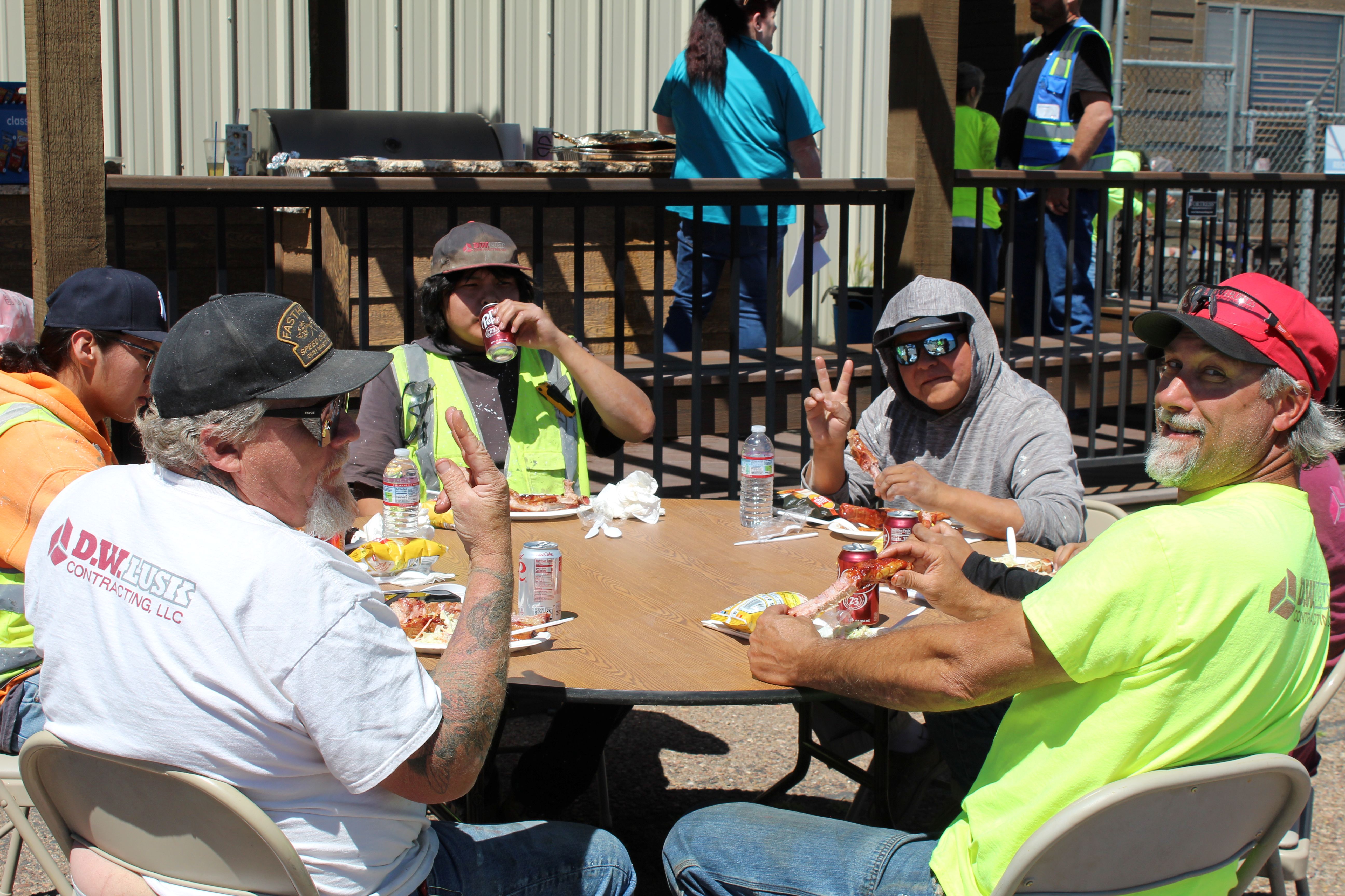Group of people seated around a round outdoor table eating a meal. Plates of food, canned drinks, and water bottles are visible on the table, with an industrial building and additional people standing in the background.