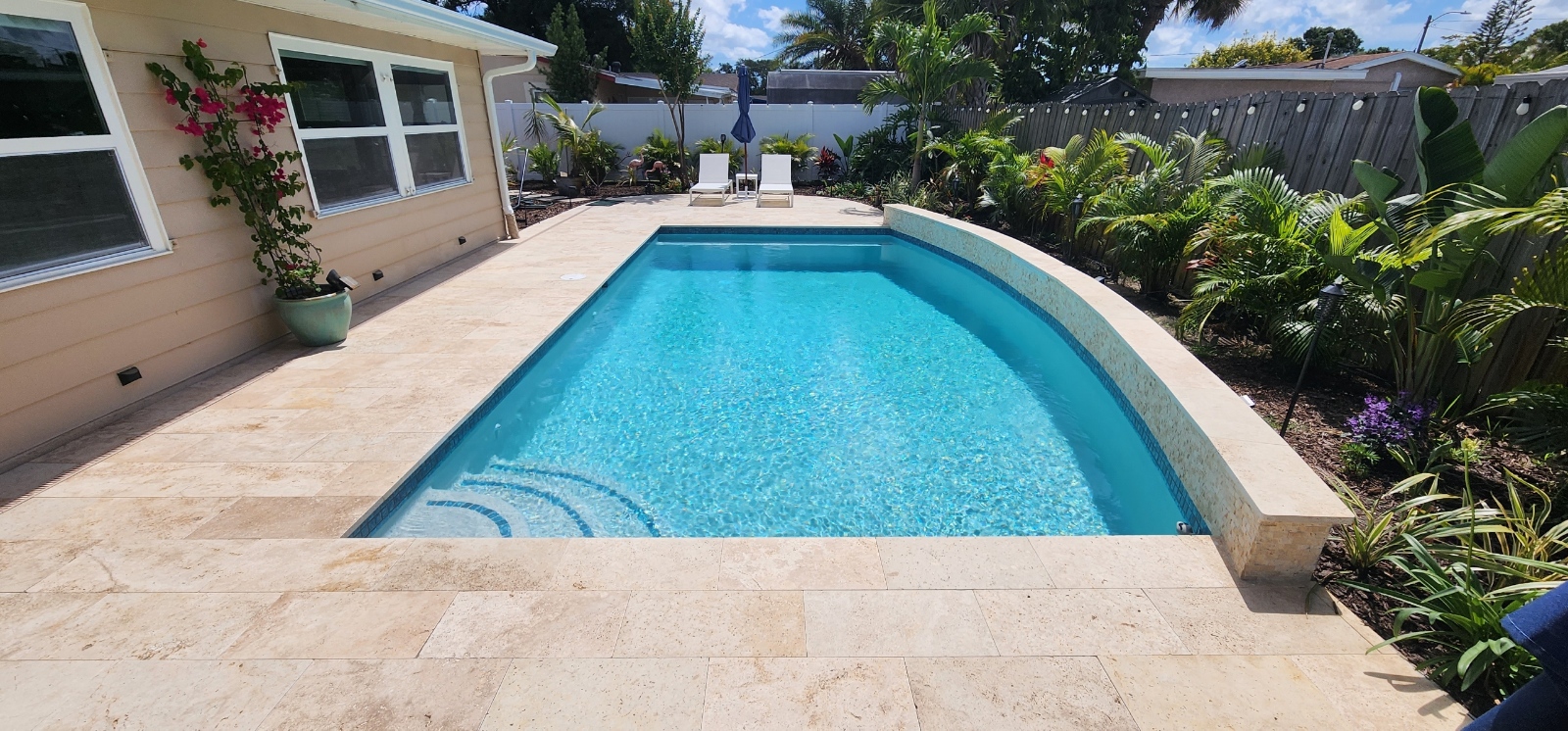 Rectangular backyard swimming pool with bright blue water, stone decking, and tropical landscaping beside the home.