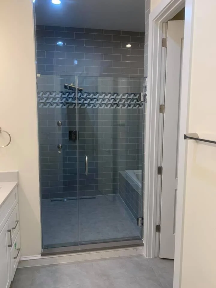 A modern bathroom featuring a spacious glass-enclosed shower with gray subway tile walls and a decorative geometric tile band. Inside the shower, a showerhead and controls are visible, along with a bathtub. To the right, a white door with silver hardware is slightly ajar, revealing a glimpse of the room beyond. The bathroom also includes a white vanity with drawers and a silver towel rack on the wall.