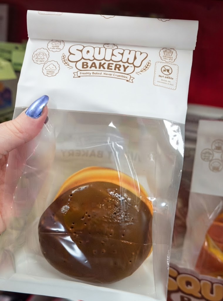 A hand holding a new Squishy Bakery toy packaged in a realistic bakery bag, showcasing an adorable, soft, and hyper-realistic pastry squishy design on store shelves.