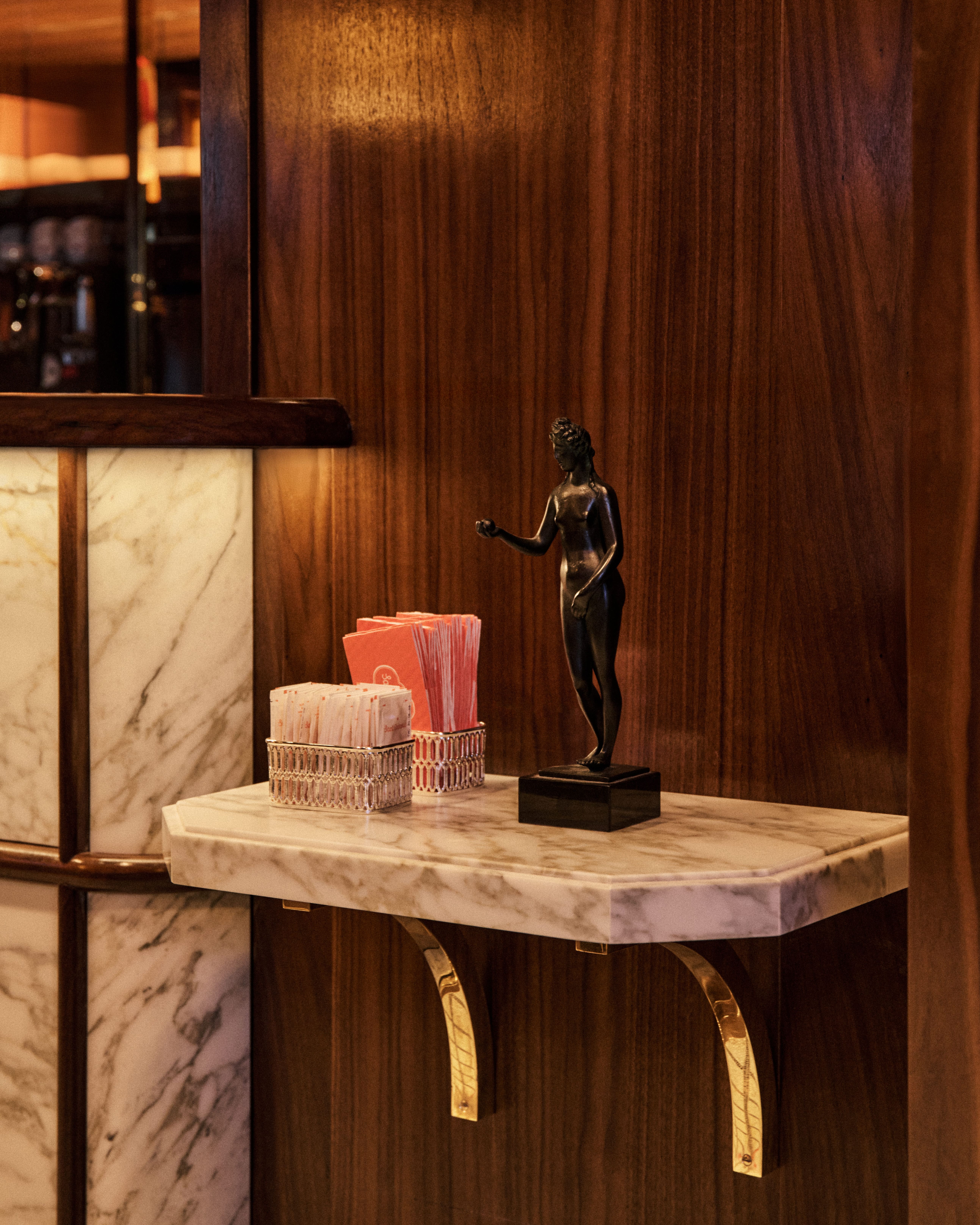 Small bronze nude figurine on a marble shelf with brass brackets, set against warm wood paneling beside the pastry counter.
