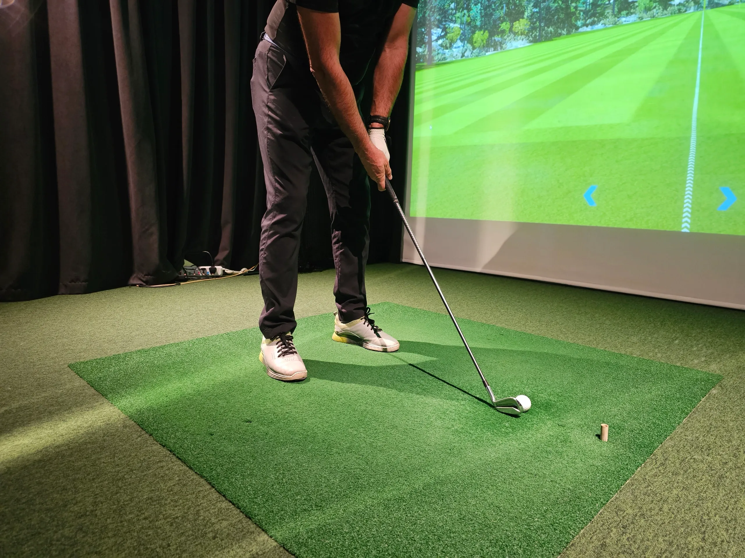 A man is about to hit a golf ball with a club on an indoor golf simulator. He is standing on a green turf mat, dressed in dark pants and a dark t-shirt. A large projection screen behind him displays a virtual golf course with trees and a fairway. The simulator includes blue arrows on the screen, suggesting interactive elements. The scene is lit, with dark curtains visible to the left.