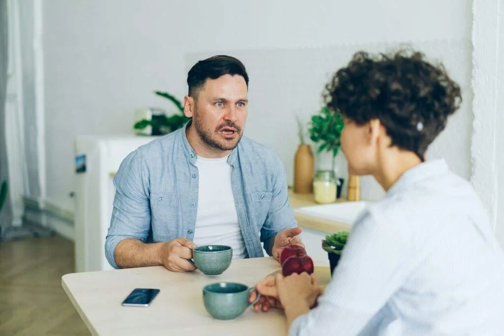 A man with a beard and short hair is sitting at a table, holding a mug and talking intensely to a person whose back is to the camera. He is wearing a light blue unbuttoned shirt over a white t-shirt. The other person has curly dark hair and is wearing a light blue collared shirt. They are also holding a mug and have two red apples in their hand. A smartphone is lying on the table. In the background, a white refrigerator and some plants are visible.
