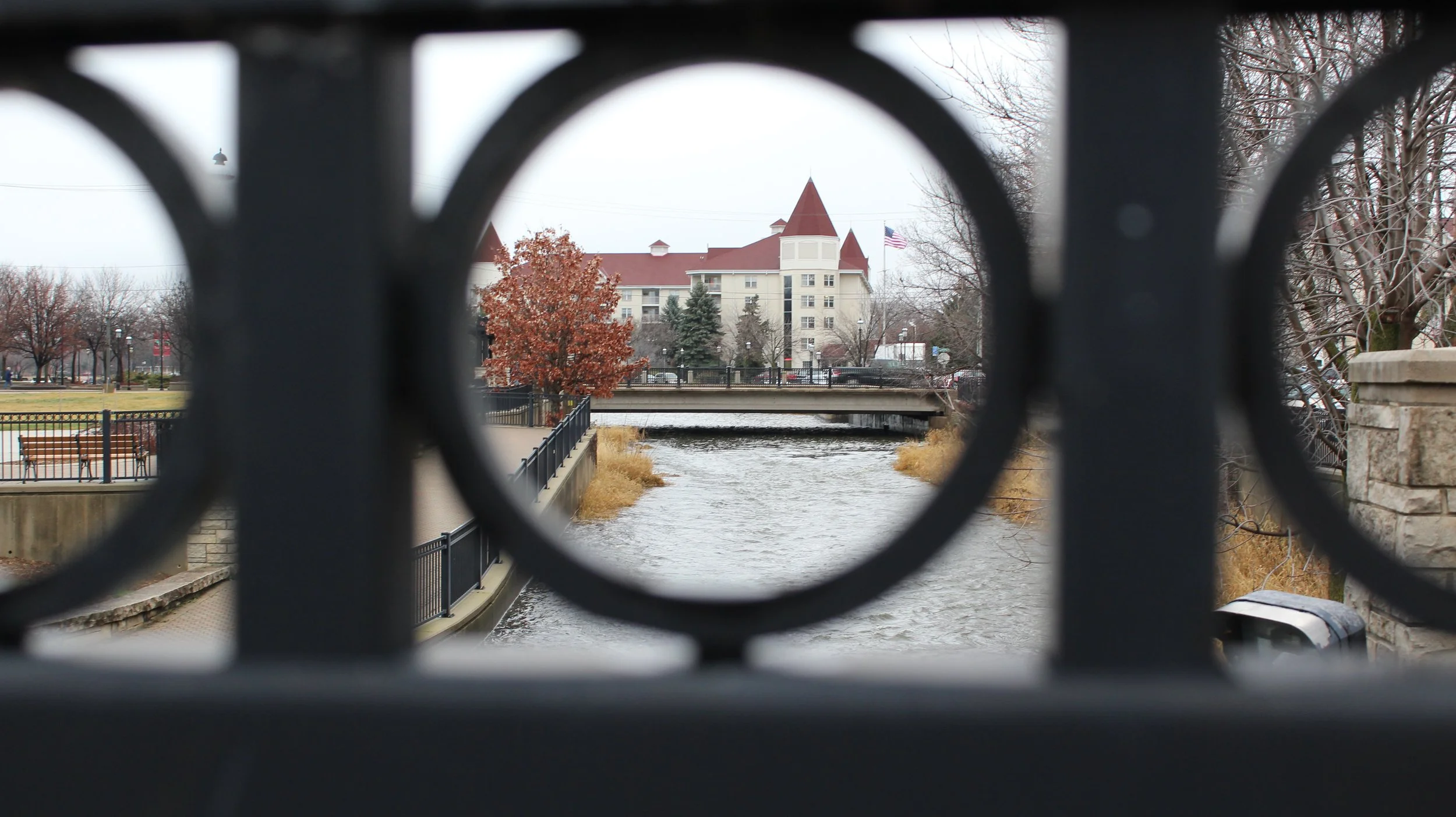 A medium shot captures a view of a building and a bridge over a river through a dark metal fence with circular openings. The building has red roofs and a light-colored facade, with an American flag flying near its top. A tree with autumnal foliage stands to the left of the building. The river flows with ripples, and dry grass grows along its banks. A walkway with a black railing runs along the river. Bare trees and a grassy area with a bench are visible in the background. The sky is overcast.