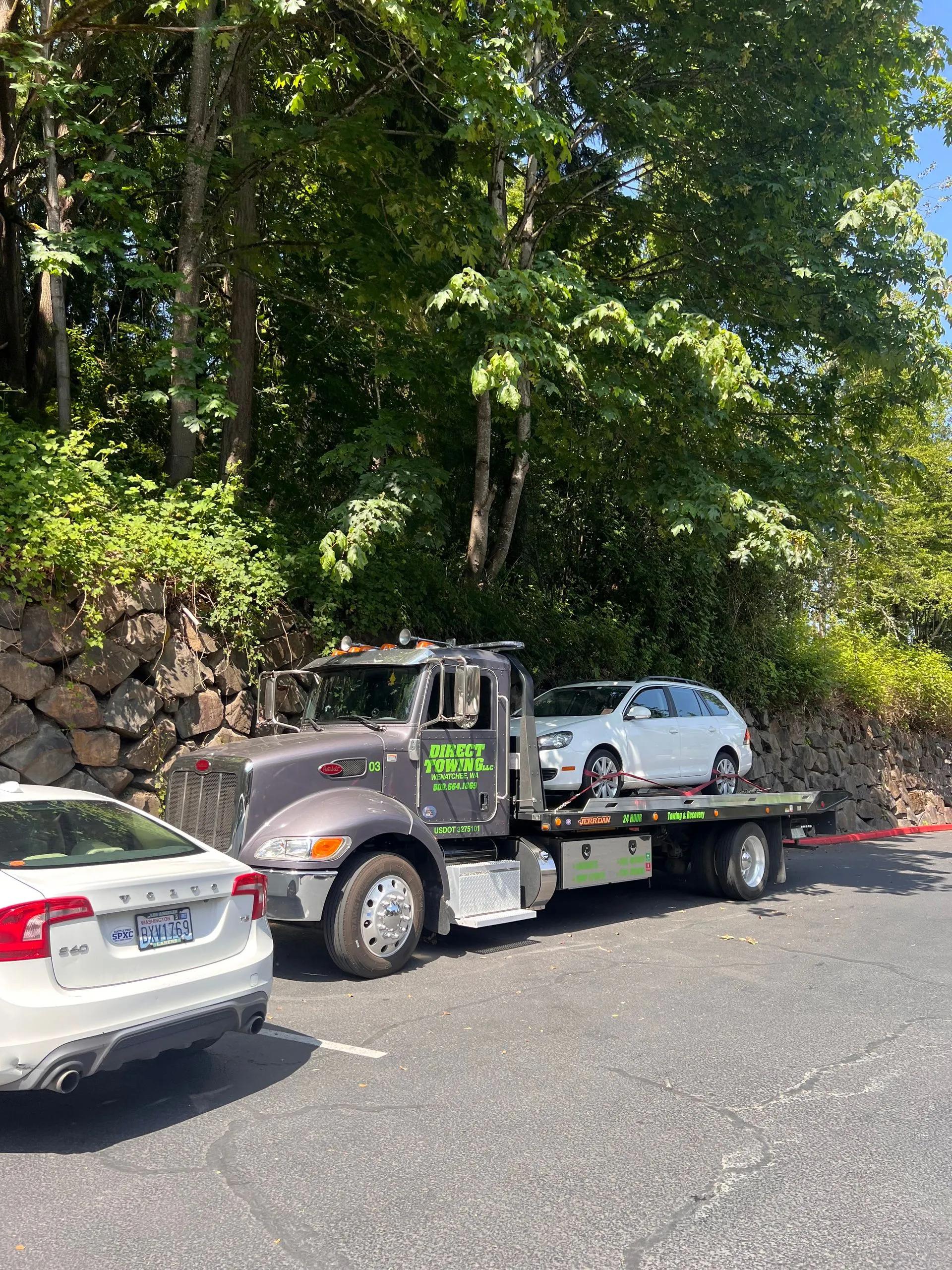 Direct Towing flatbed truck transporting a white vehicle along a roadside location surrounded by trees and stone retaining walls, demonstrating secure vehicle transportation and professional towing services.